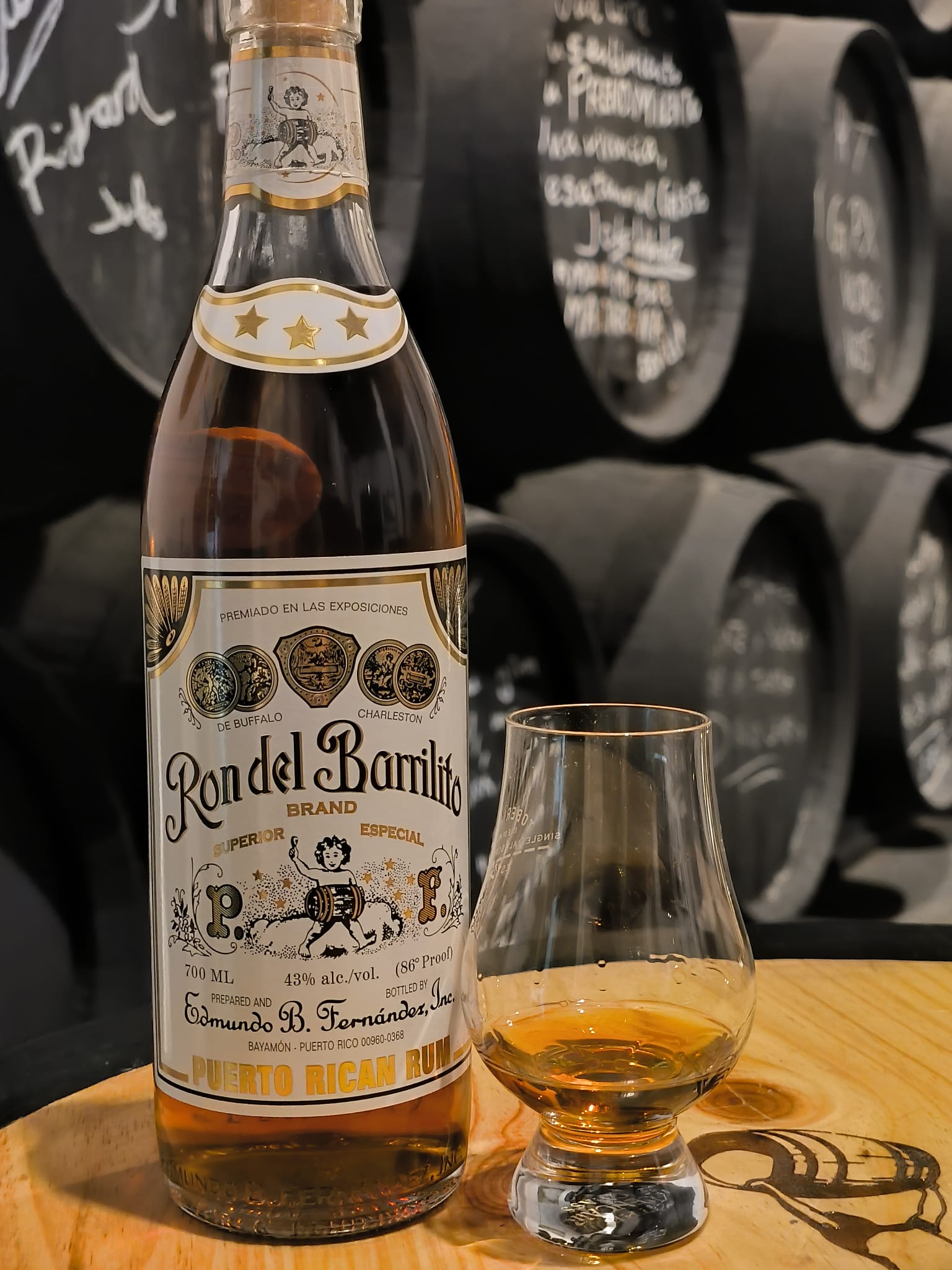 Botella de Ron del Barrilito junto a una copa y barricas